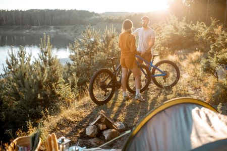 Young couple standing with mountain bicycles at the campsite, traveling in the forest near the lake on the sunset